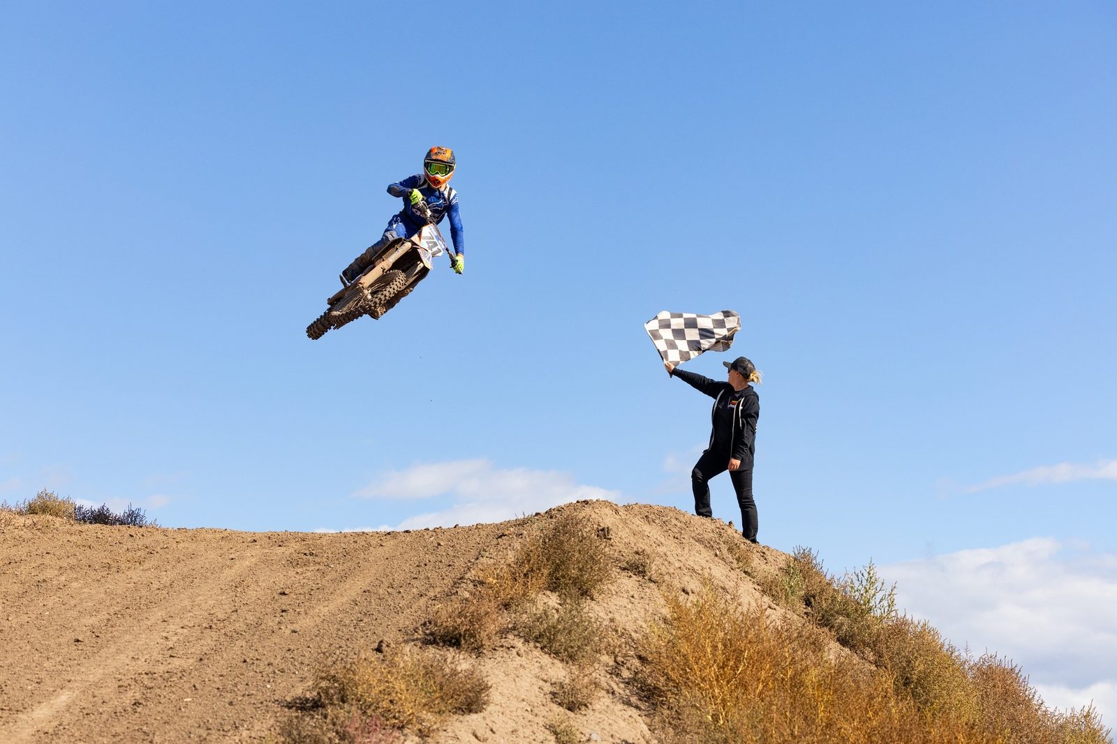 A dirt bike rider in the air over a jump at Dry Lake Motocross Park while a flagger waves a checkered flag from the hilltop