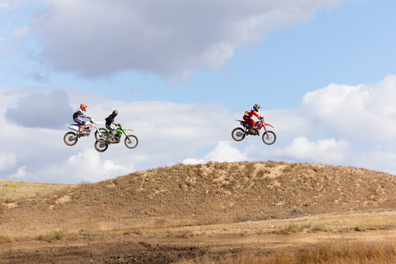 Three motocross riders in the air over a jump at Dry Lake Motocross Park against a big Colorado sky