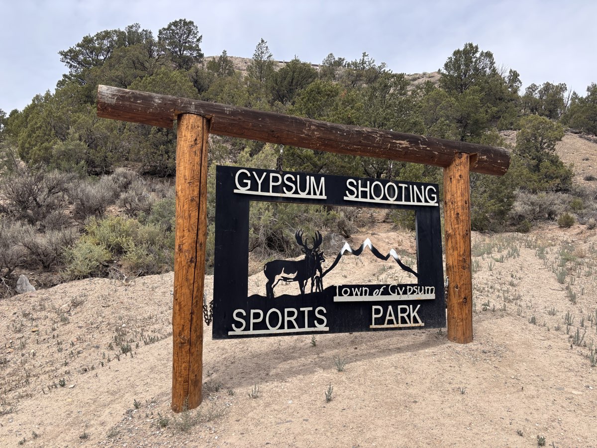 The timber and steel entrance sign at the Town of Gypsum Shooting Sports Park, cut with a mule deer buck and mountain skyline, set in sagebrush and pinyon juniper