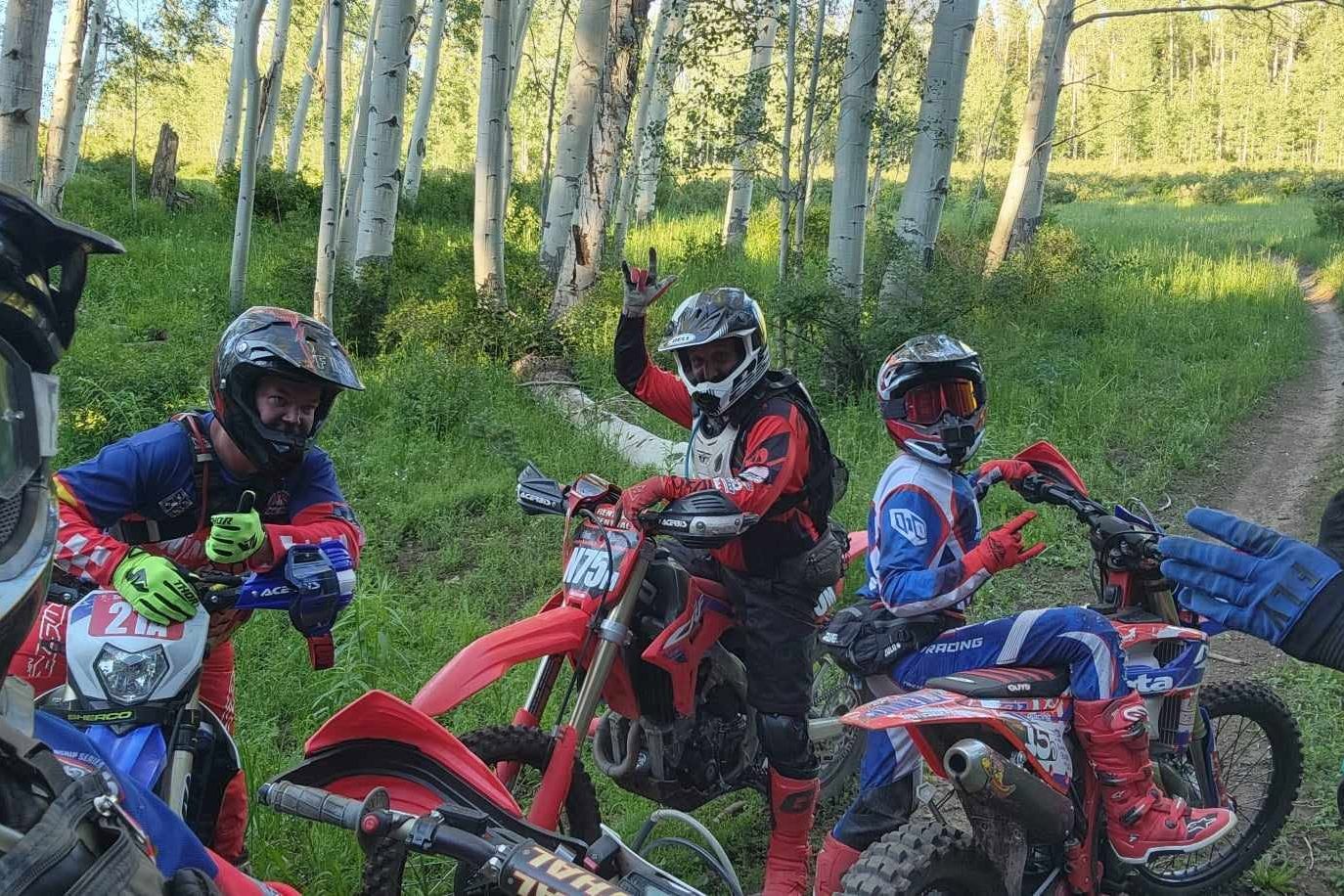 Three dirt bike riders in full gear pausing among aspen trees on a Hardscrabble trail near Spring Creek, one waving at the camera